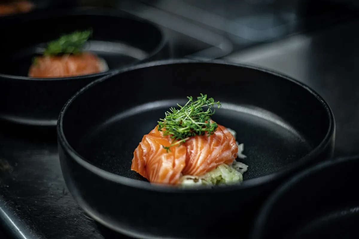 Elegantes Gericht mit rosa Lachsfilet und frischen Kr&auml;utern, angerichtet auf schwarzem Teller in gehobener K&uuml;che.