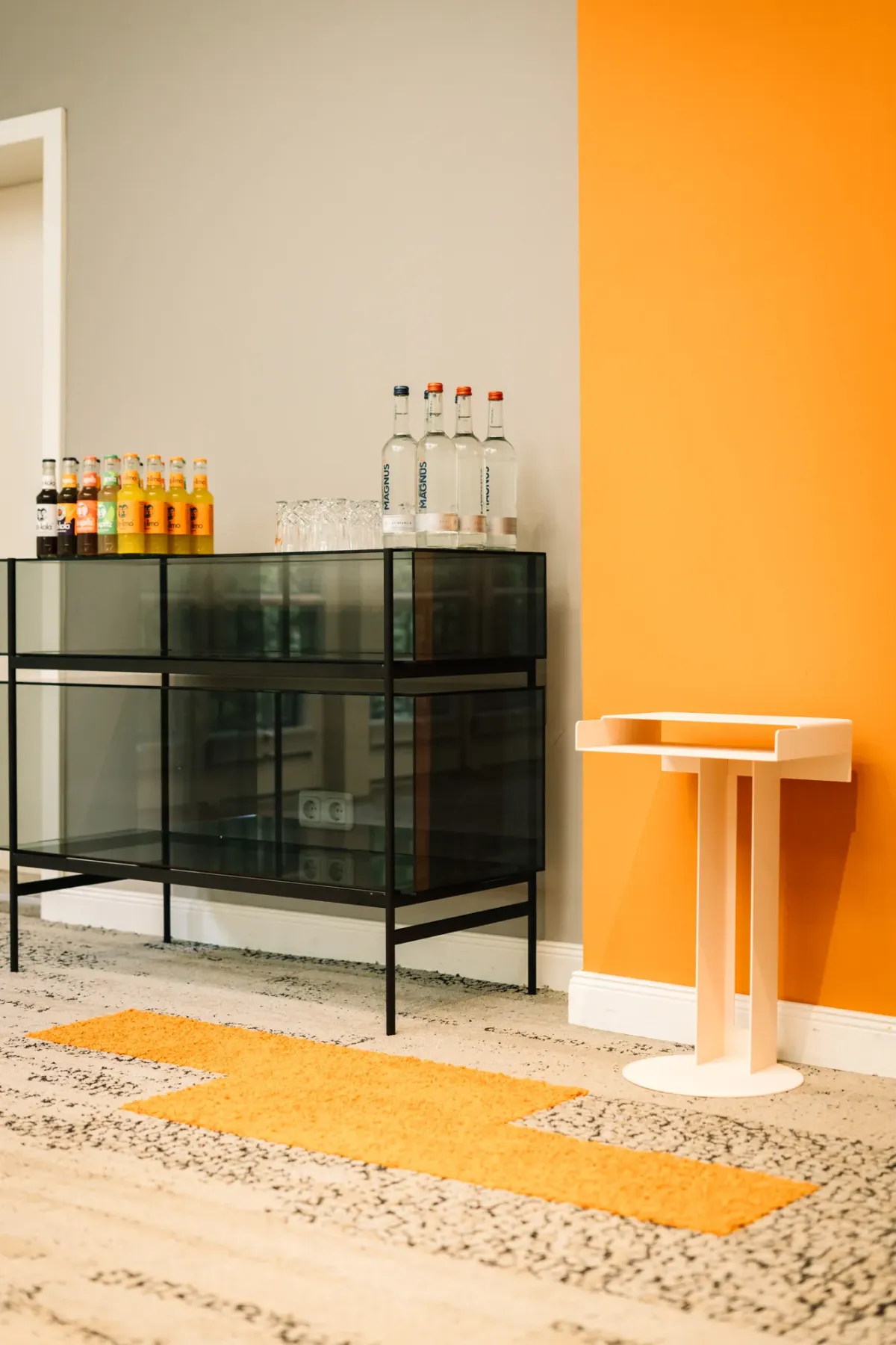 Moderner Tagungsraum mit orangefarbener Wand, schwarzem Metallregal mit Getr&auml;nkeflaschen.