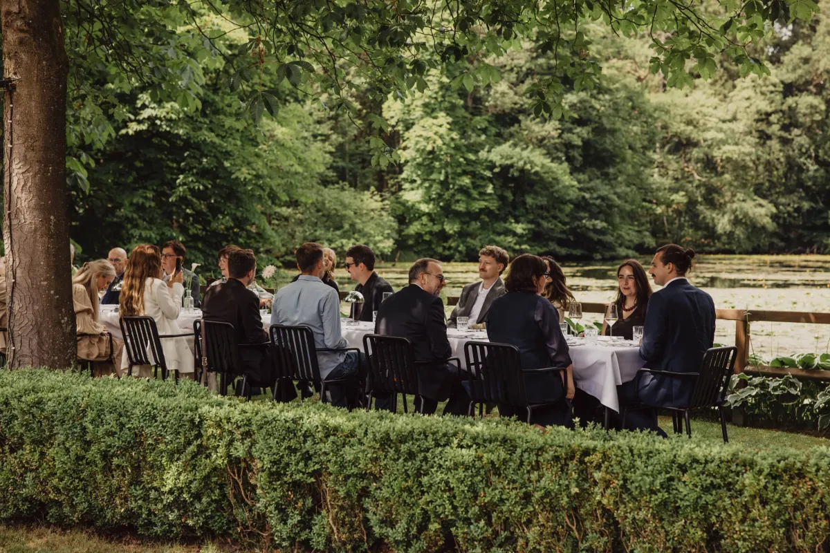 Elegante Hochzeitsgesellschaft sitzt an gedecktem Tisch im Freien unter alten B&auml;umen an einem See.