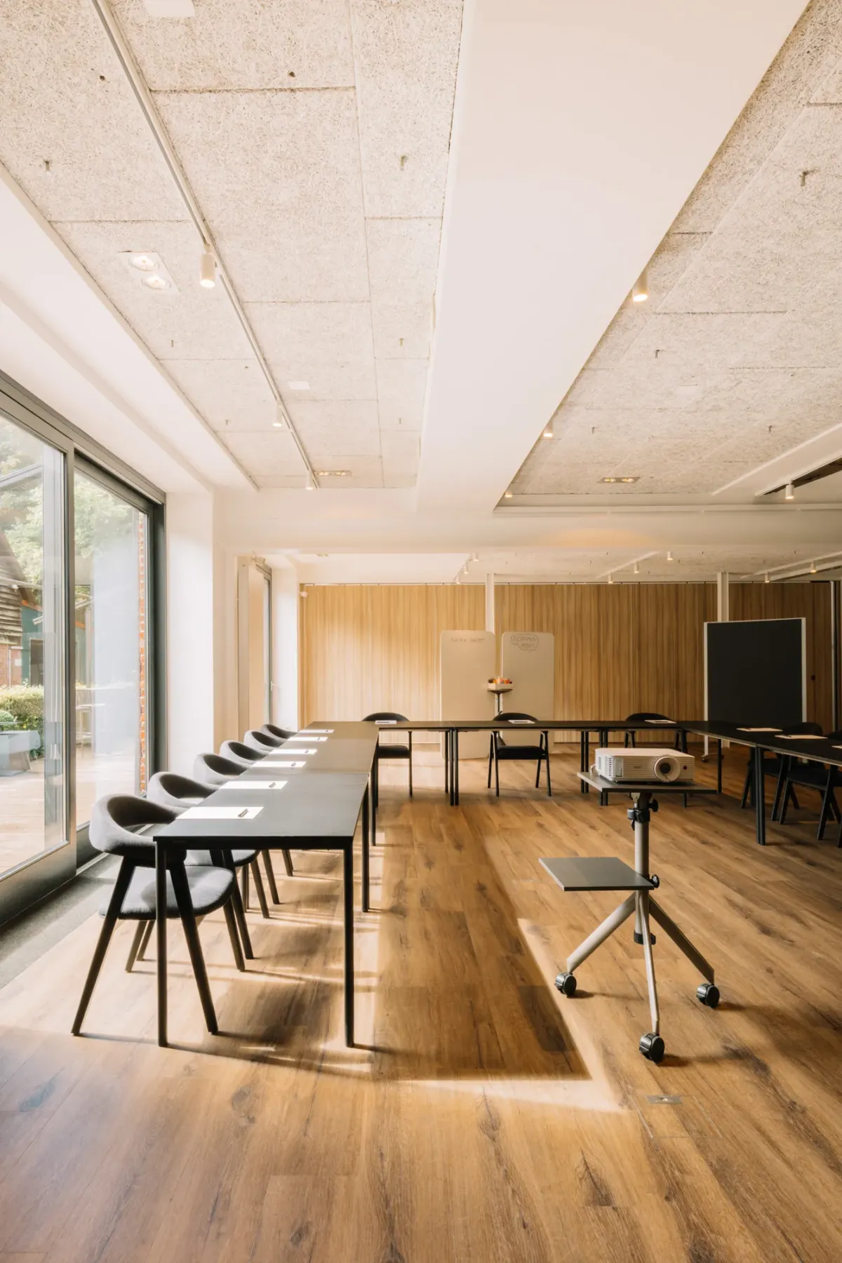 Moderner Veranstaltungsraum mit Holzparkett, hellen Decken, gro&szlig;en Fenstern und flexibel arrangierten schwarzen St&uuml;hlen sowie Tischen f&uuml;r Events und Meetings.