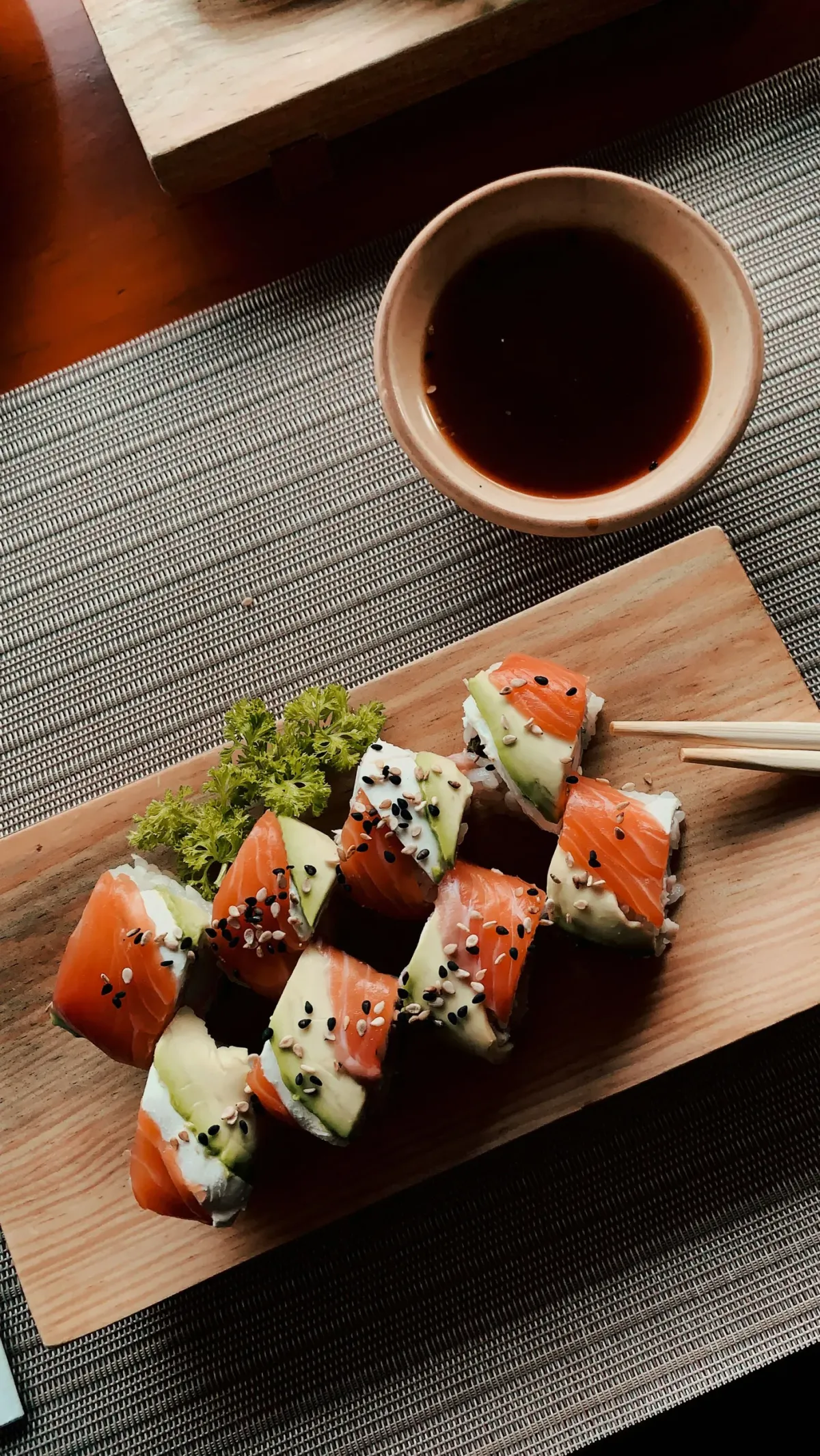 Sushi-Rollen auf Holzbrett serviert mit Sojasauce in wei&szlig;er Schale, arrangiert auf grauem Tischl&auml;ufer.