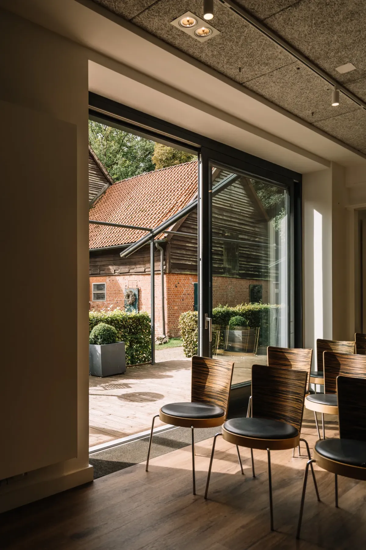 Moderner Veranstaltungsraum mit gro&szlig;en Glasschiebet&uuml;ren, die zu einer Terrasse mit Sitzbereichen f&uuml;hren. Minimalistische Einrichtung mit Metallst&uuml;hlen und Blick auf das historische Ziegelsteingeb&auml;ude und die umgebende Natur.