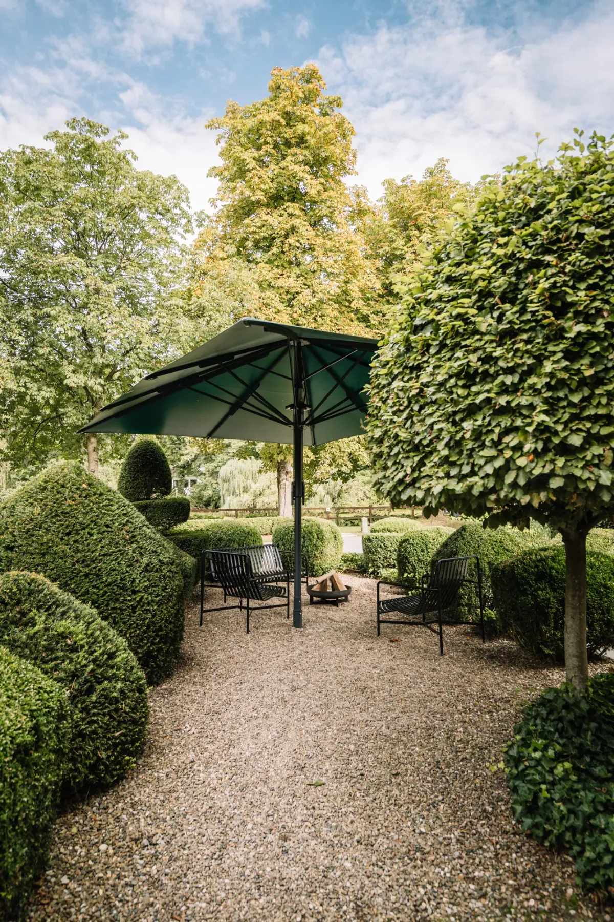 Ein Gartenplatz mit schwarzem Sonnenschirm und Sitzm&ouml;beln, umgeben von gr&uuml;nen Hecken und hohen B&auml;umen unter blauem Himmel.