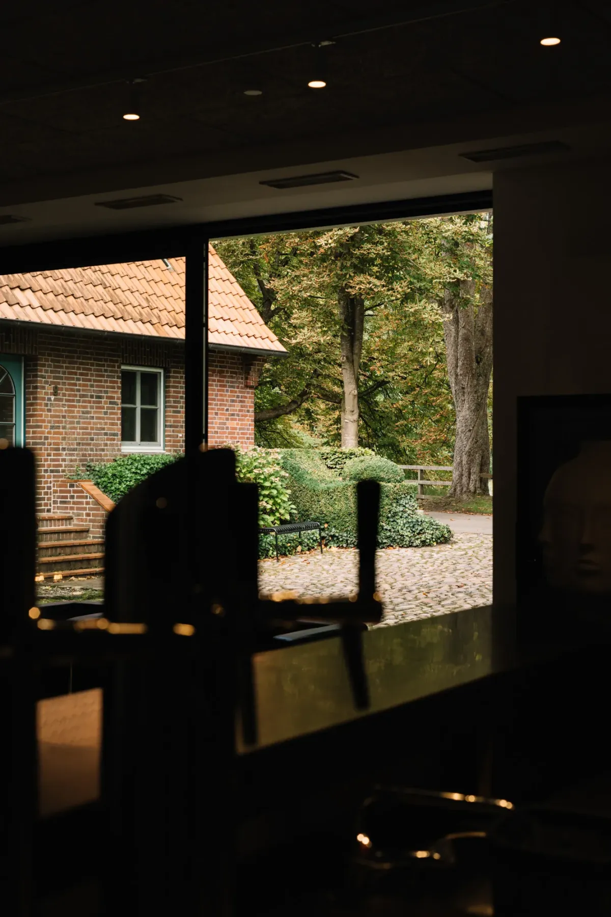 Blick durch eine offene T&uuml;r auf einen Innenhof mit Backsteingeb&auml;ude, Kiesweg und gr&uuml;nen B&auml;umen im Hintergrund.