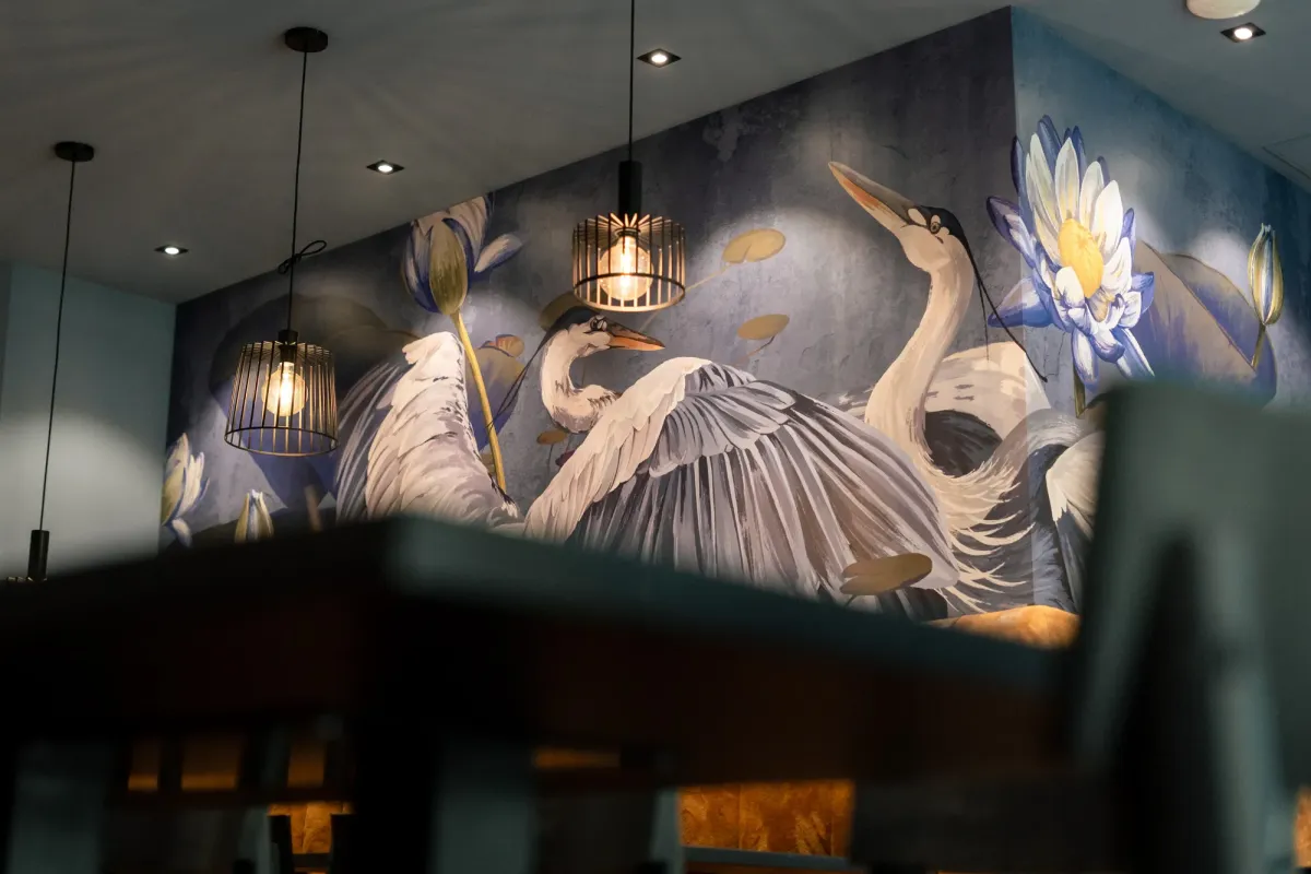 Elegante Wandmalerei mit wei&szlig;en Reihern und Wasserlilien an dunkler Wand, illuminiert durch h&auml;ngende Lampen in gehobener Eventlocation.