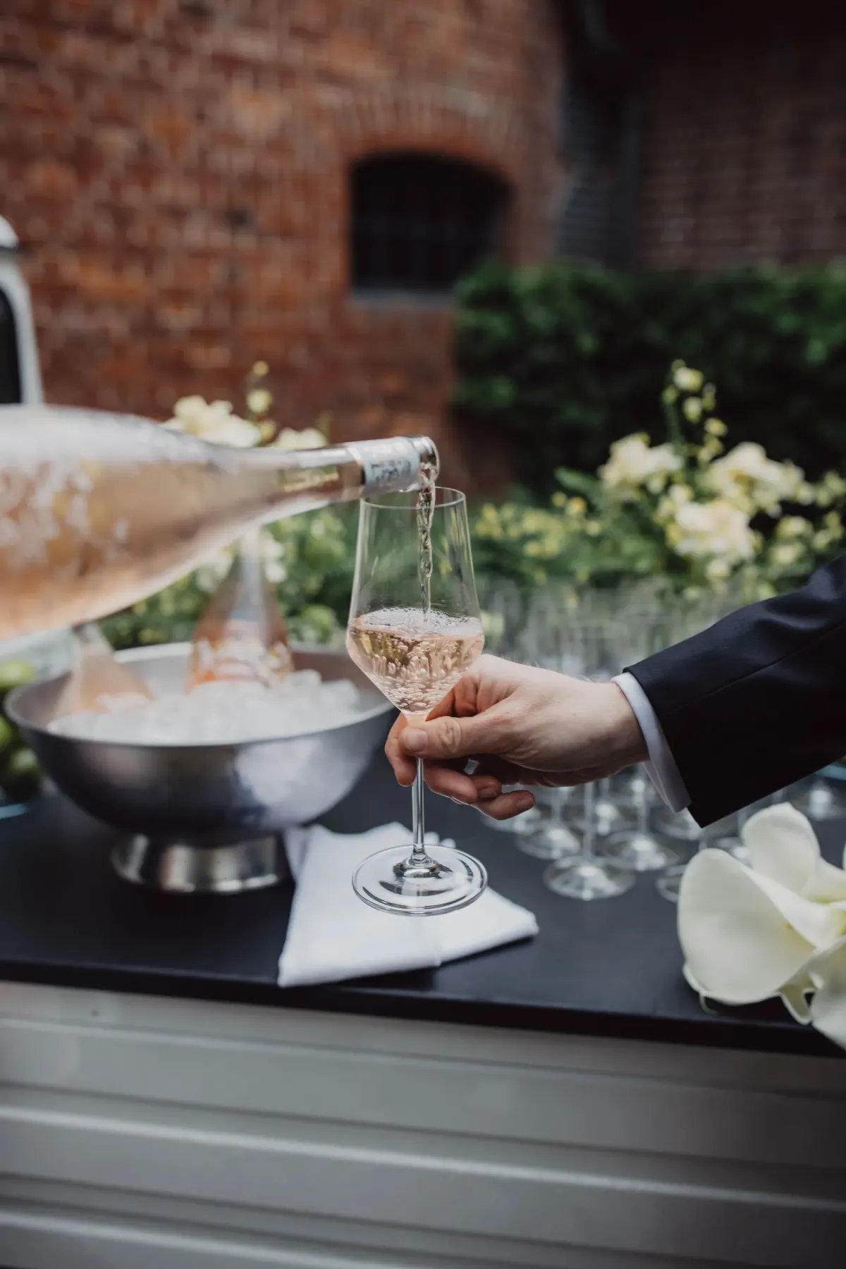 Person in schwarzem Anzug schenkt Ros&eacute;-Wein in ein Glas ein. Im Hintergrund eine Sch&uuml;ssel mit Speisen und Blumendekorationen auf einer schwarzen Tafel. Ziegelmauer und Gr&uuml;n im unscharfen Hintergrund.