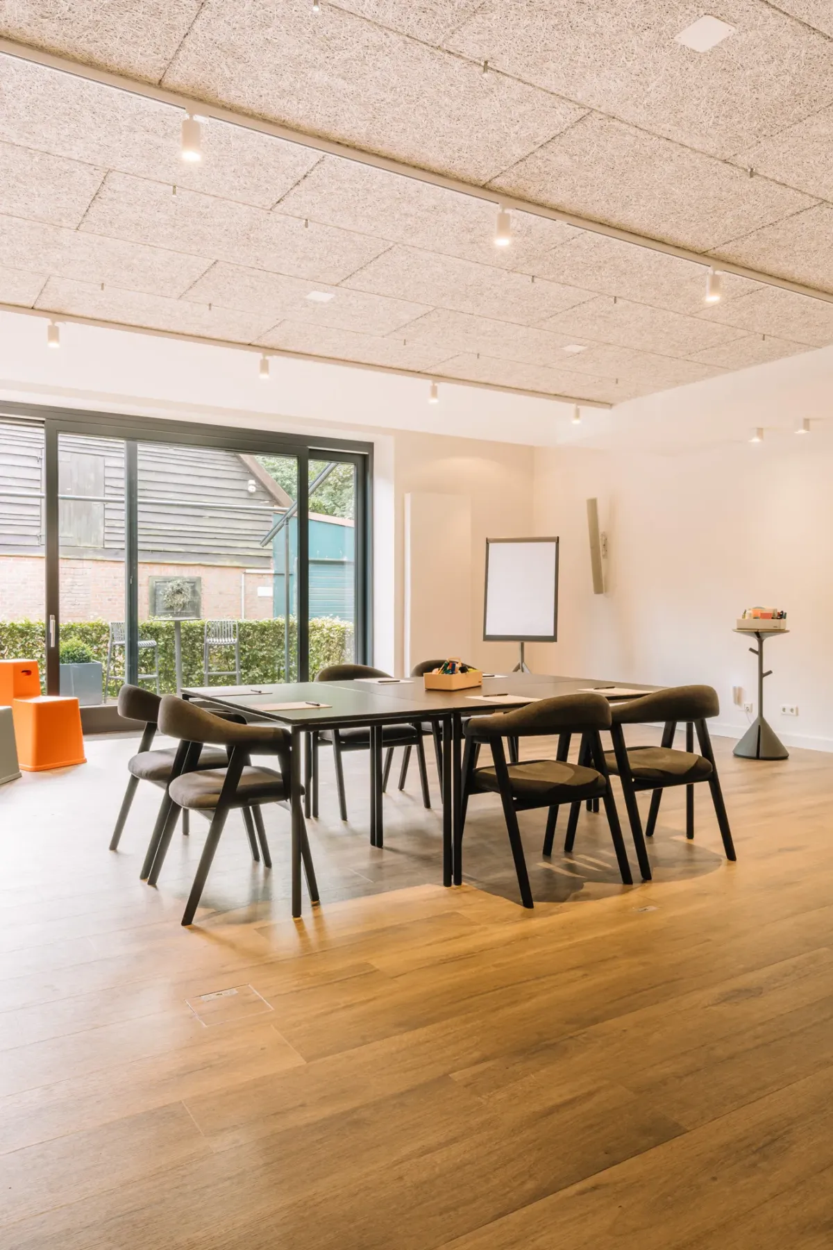 Moderner Meetingraum mit Holzdecke, gro&szlig;en schwarzen Tischen, sechs St&uuml;hlen und bodentiefem Fenster zur Terrasse mit Gartenblick.