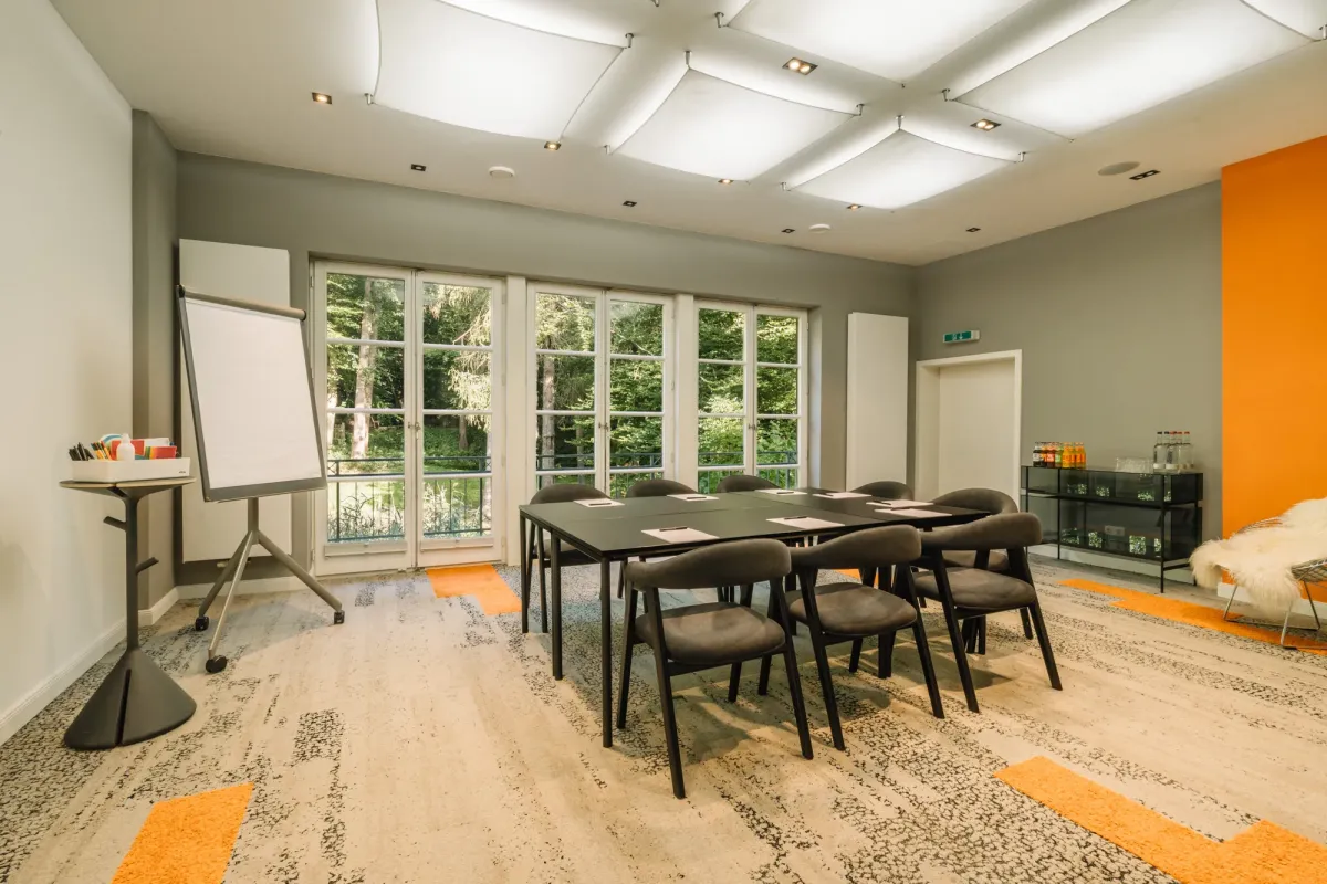 Moderner Seminarraum mit gro&szlig;z&uuml;gigen Fenstern, schwarzem Konferenztisch, ergonomischen St&uuml;hlen, Flipchart und orange-grauen Designelementen.