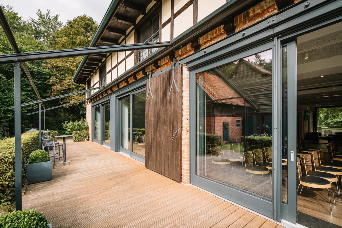 Moderne Terrasse mit gro&szlig;fl&auml;chigen Glasschiebet&uuml;ren, Holzdecking und Stahlkonstruktion, umgeben von gr&uuml;nem Garten und historischem Fachwerkgeb&auml;ude.